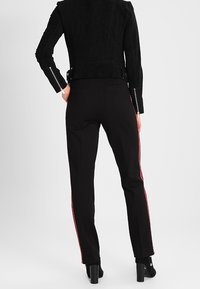 Osoba v černých přiléhavých kalhotách s červenými bočními pruhy, černé bundě se zipovými detaily na rukávech a černých kotníkových botách na podpatku.