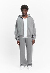 Gray zip-up hoodie with a front pocket and matching sweatpants. Soft fabric, relaxed fit, and white T-shirt underneath, complemented by white sneakers.