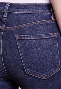 Tummansininen farkut, joissa on tiukka muotoilu, näkyvät ompeluakkentit ja tavallinen takatasku. Sujuva pinta.