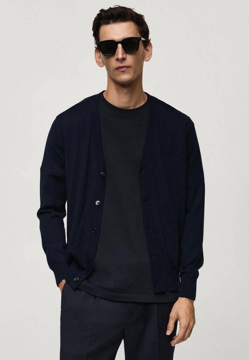 Marinblå cardigan i stickad tyg, med V-hals, mörk knappstängning och långa ärmar, lager på lager över en mörk t-shirt.