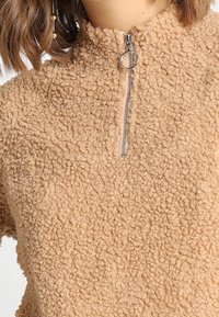 Polaire beige avec un col montant et une fermeture éclair à moitié. Présente un tissu doux et texturé avec un motif en boucles.