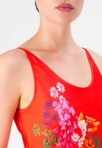 Mujer vistiendo una blusa sin mangas roja con un patrón floral en la parte delantera en tonos rosa, púrpura, blanco y naranja sobre un fondo liso.