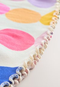 Couverture en tissu doux avec des pois multicolores en rose, orange, violet et bleu. Elle présente une bordure froncée crème le long du bord.