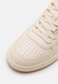 Crème-kleurige sneakers met perforaties, dikke witte veters en rubberen zool, getoond vanuit een close-up hoek.