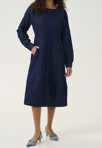 Jeansklänning med långa ärmar, rund halsringning och sidfickor. Mörkblå färg med en subtil struktur och knälång fåll.