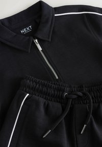 Fekete cipzáros polo ing és elasztikus derekú fekete nadrág fehér díszítéssel és zsinóros részletekkel, laposan elhelyezve.