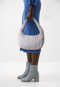 Hellgraue gepolsterte Handtasche mit geschwungener Form, ausgestattet mit einem Reißverschluss und einer weichen Textur, gehalten neben blauen Ankle-Boots und einem strukturierten Kleid.