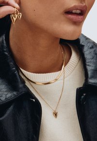 Goldene, herzförmige Ohrringe und eine passende Anhänger-Halskette, übereinander getragen über einem cremfarbenen Pullover, kombiniert mit einer schwarzen Lederjacke.