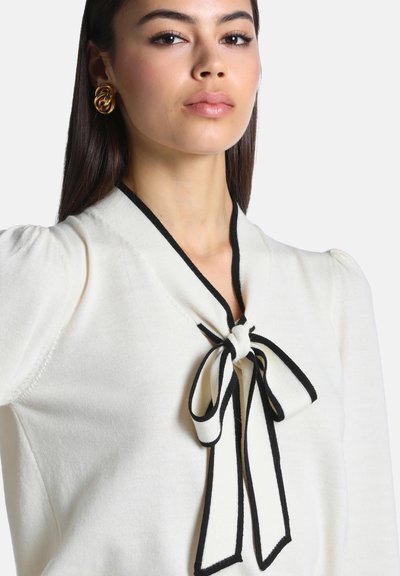 Jeune femme aux longs cheveux foncés portant des boucles d'oreilles en forme de nœud en or et un haut blanc avec un nœud bordé de noir noué au niveau de l'encolure.