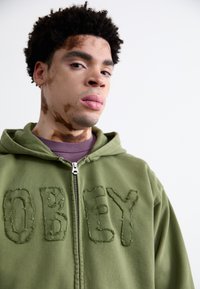 Olive green zip-up hoodie with distressed "OBEY" embroidery on the front, featuring a kangaroo pocket and a drawstring hood.