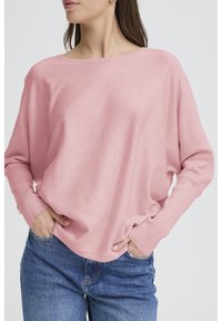 Pull surdimensionné en maille rose clair avec un col large et des poignets côtelés, associé à un jean bleu. Texture douce, coupe décontractée.