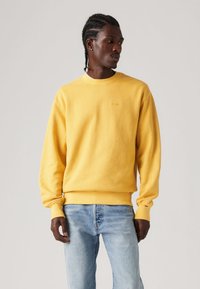 Yellow sweatshirt made of soft fabric, featuring a round neckline, ribbed cuffs, and a subtle logo embossed on the chest. Paired with light blue jeans.