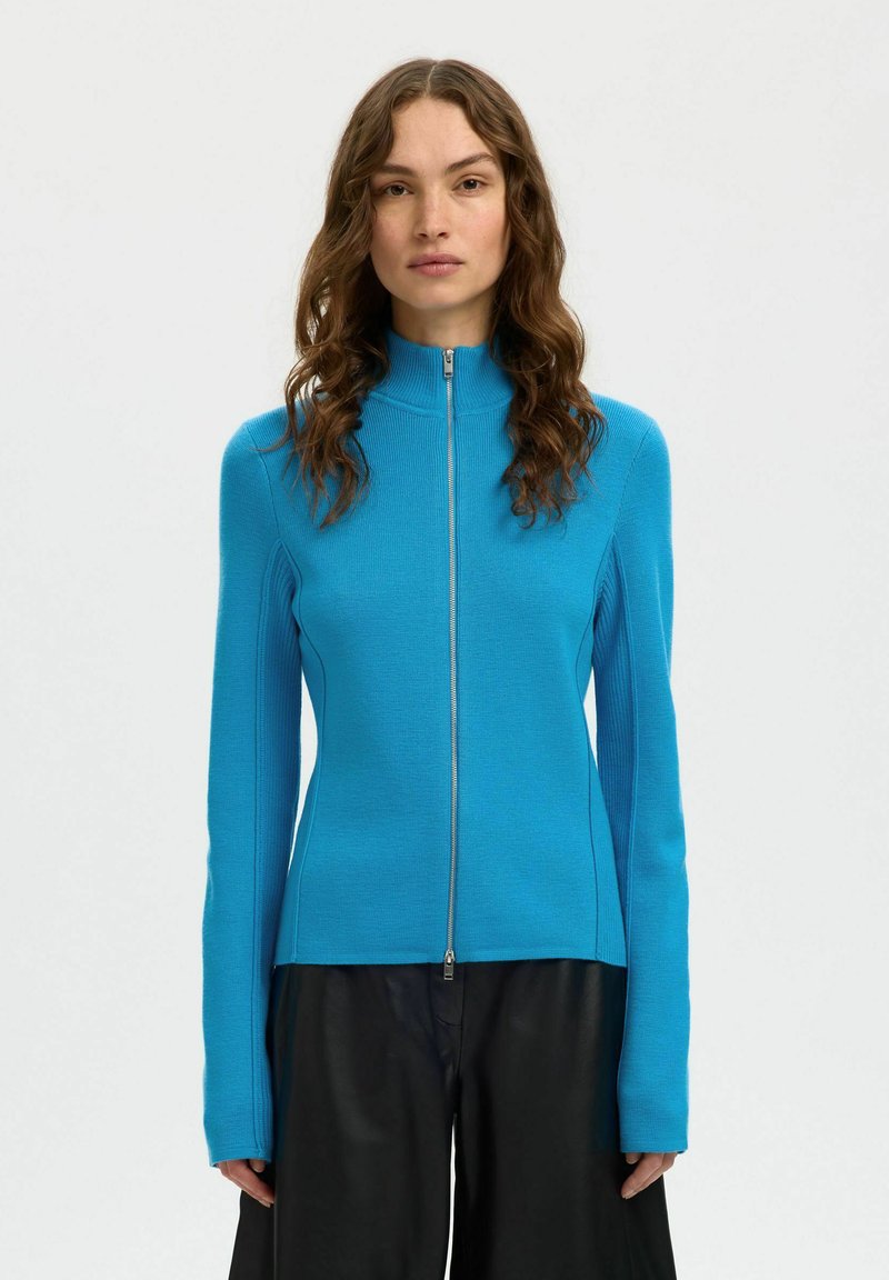 Vrouw met golvend bruin haar, gekleed in een felblauwe sweater met rits en zwarte wijde broek, staand tegen een eenvoudige lichte achtergrond.