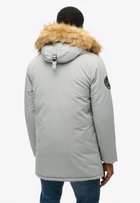 Parka aislante gris con capucha con borde de pelaje, correa de cuello ajustable y un parche circular en la manga izquierda. Textura suave, diseño hasta la rodilla.