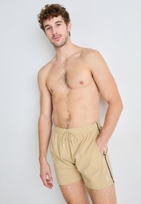 Shorts de bain beiges avec une taille élastique, fermeture à cordon, et rayures sur les côtés. Fabriqués en matériau léger.