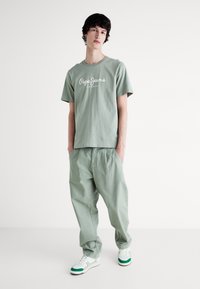 Joven de pie con las manos en los bolsillos, vistiendo una camiseta sage green de Pepe Jeans, pantalones sueltos a juego y zapatillas blancas con detalles verdes.