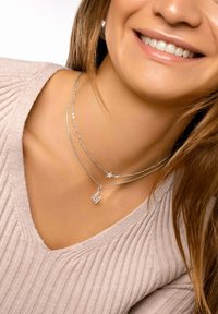 Colliers superposés en argent avec une petite étoile et un pendentif "M", portés sur un haut en maille légère, sur un fond lisse et neutre.
