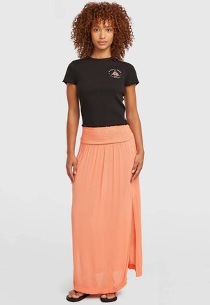 Femme aux cheveux bouclés portant un t-shirt noir ajusté et une jupe maxi corail taille haute avec une fente latérale, debout les bras détendus.