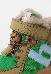 Zapato infantil verde y beige con cordones elásticos marrones, forro de piel, correa de Velcro etiquetada "bisgaard" y detalle del logo azul en el lateral.