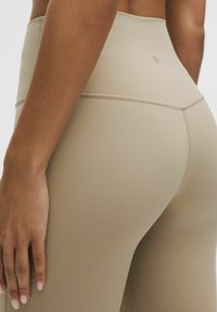 Béžové legíny s vysokým pásom a hladkou, elastickou textúrou. Obsahujú minimálne švy a jemné logo blízko pásu.