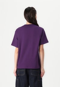 Vrouw met lang, donker haar staat met haar rug naar ons gekeerd, in een eenvoudige paarse t-shirt met korte mouwen en donkere denim broek met grote zakken.