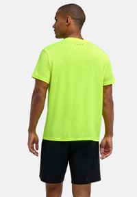 Mężczyzna ubrany w jasny żółty sportowy t-shirt z krótkim rękawem i czarne spodenki, stojący tyłem na jednolitym białym tle.