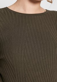 Oliwkowo zielony prążkowany sweter z fakturowanym wzorem z pionowymi liniami. Widok z bliska pokazuje dzianinę oraz okrągły dekolt.