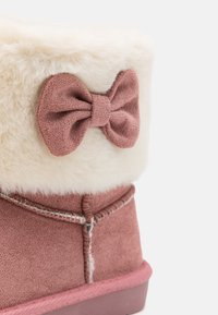 Botte en suede rose avec un revers en fausse fourrure blanche, accentuée par un nœud rose. Présente des détails cousus sur les côtés et une semelle texturée.