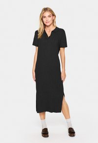 Svart poloklänning i knälång längd med korta ärmar, ribbad textur, sidoklyftor och en klassisk krage. Stylad med bruna skor och grå strumpor.