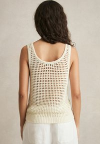 Ljusbeige stickad linne med öppen vävdesign, ribbad nederkant och breda ärmhål som avslöjar en texturerad rygg.