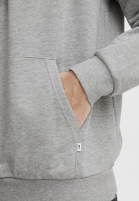 Sudadera gris con un bolsillo delantero, puños acanalados y una pequeña etiqueta con el logo en el lateral. La tela es suave y tiene una textura agradable.