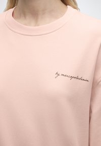 Ροζ ανοιχτό ροζ φούτερ με στρογγυλή λαιμόκοψη και μικρό μαύρο καλλιγραφικό κείμενο «by marcopolodenim» στην αριστερή πλευρά του στήθους.