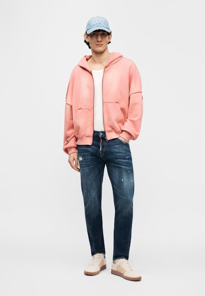 Ροζ φούτερ με κουκούλα, λευκό αμάνικο μπλουζάκι, σκούρο μπλε τζιν με σκισίματα και μπεζ αθλητικά παπούτσια. Γαλάζιο καπέλο με το κείμενο "Riot". Casual στυλ.