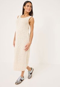 Abito lungo senza maniche beige in maglia con scollo tondo; indossato con sneakers grigie e blu con accenti metallici e dettagli texturizzati.