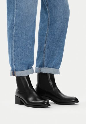 Czarne skórzane botki na niskim szerokim obcasie, noszone z podwiniętymi niebieskimi jeansami, na białym tle.