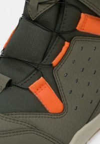 Gros plan sur une sneaker vert olive avec un tissu texturé, des onglets de couture orange, des lacets en fil noir et un détail de panneaux latéraux perforés.