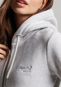 Sudadera de cremalleras en gris, hecha de una tela suave, con un bolsillo tipo canguro. Presenta un logotipo bordado en el pecho y cordones en la capucha.