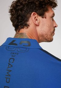 Camisa polo de manga corta azul con cuello negro, gran texto impreso "CAMP" y "63" en la parte posterior, con tejido texturizado.