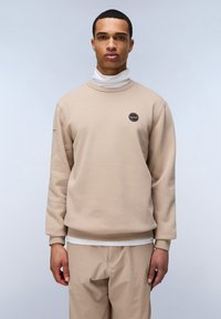Felpa beige con collo rotondo, maniche lunghe e un piccolo logo circolare sul petto. Indossata sopra un dolcevita bianco. Texture morbida.