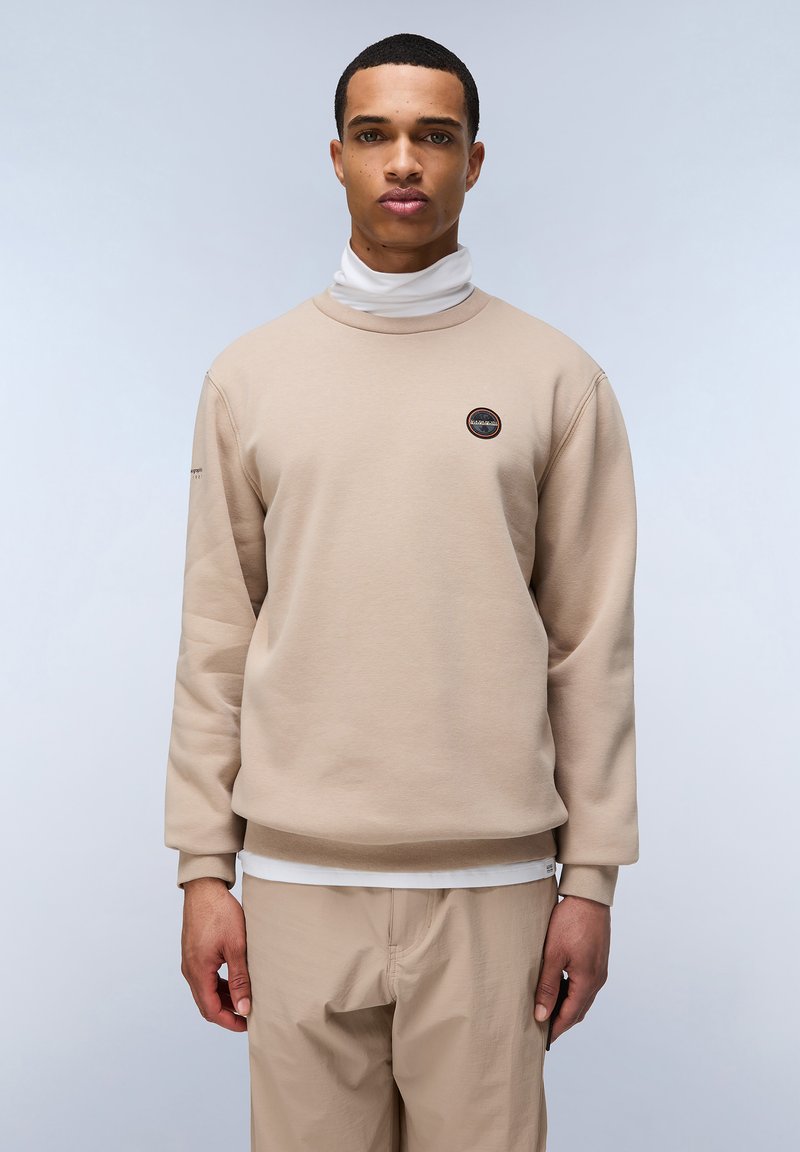Felpa beige con collo rotondo, maniche lunghe e un piccolo logo circolare sul petto. Indossata sopra un dolcevita bianco. Texture morbida.