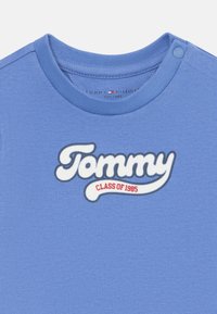 Blå bomull T-skjorte med rund hals og trykknapp. Har en stor hvit og rød logo med teksten "Tommy CLASS OF 1985".