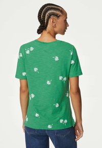 Camiseta de algodón verde con bordado floral blanco, corte relajado, mangas cortas y cuello redondo. La vista trasera muestra el patrón y la textura.