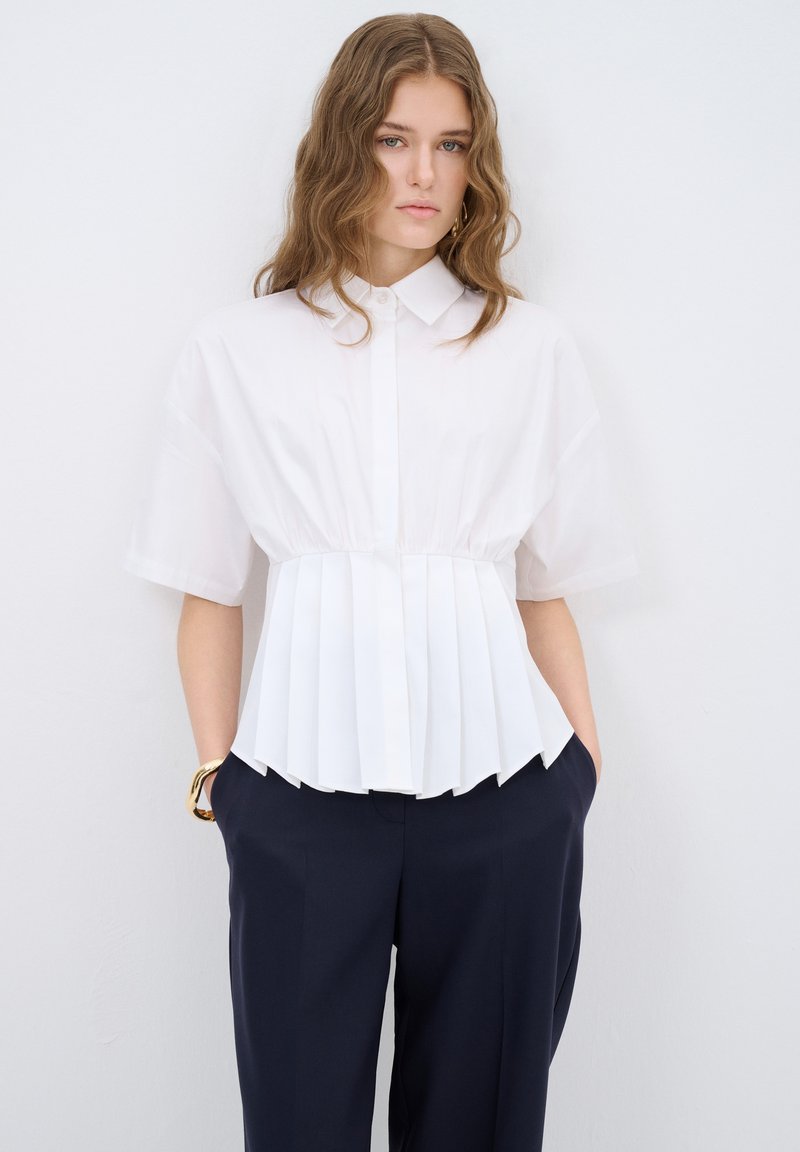 Blusa blanca de manga corta con diseño plisado en la cintura, con cuello y cierre de botones al frente. Combinada con pantalones azul marino ajustados.