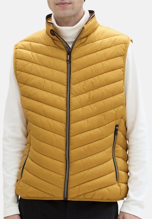 Gul quiltet vest med høj krave, frontlynlås og to sidelommer. Har chevron-syninger og en kontrastkant ved kraven.