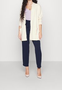 Ljusbeige cardigan med öppen front och två fickor, kombinerad med en ljuslila topp och marinblå tapered byxor. Modellen bär nudefärgade klackar.