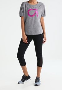 Szara koszulka sportowa z różowym stylizowanym logo, połączona z czarnymi legginsami za kostki z siateczkowymi akcentami i szaro-niebieskimi trampkami.