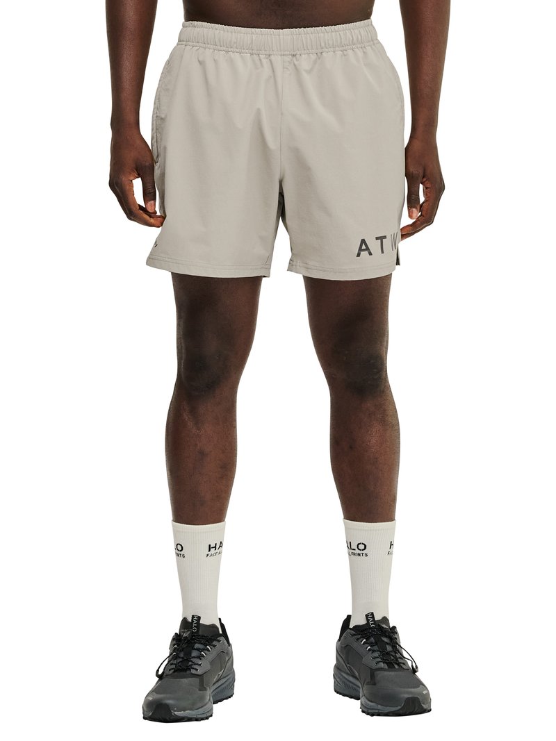 Mężczyzna w beżowych sportowych szortach z nadrukiem "ATW" na lewej udzie, białych skarpetkach z czarnym napisem oraz czarnych butach do biegania, stojący na białym tle.