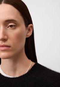 Nahaufnahme einer Person, die einen dunklen Wollpullover trägt. Sie hat glattes Haar und eine Perlenkette. Der Hautton ist hell mit subtilen Texturen.