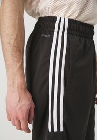 Närbild av en person som bär svarta träningsshorts med vit tre-strips design och en ljusfärgad meshskjorta.