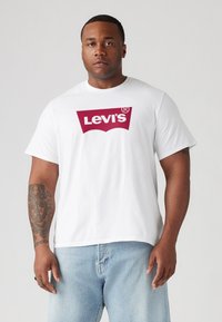 White cotton t-shirt featuring a large maroon Levi's logo on the chest. The fabric is smooth with a relaxed fit, paired with light blue jeans.